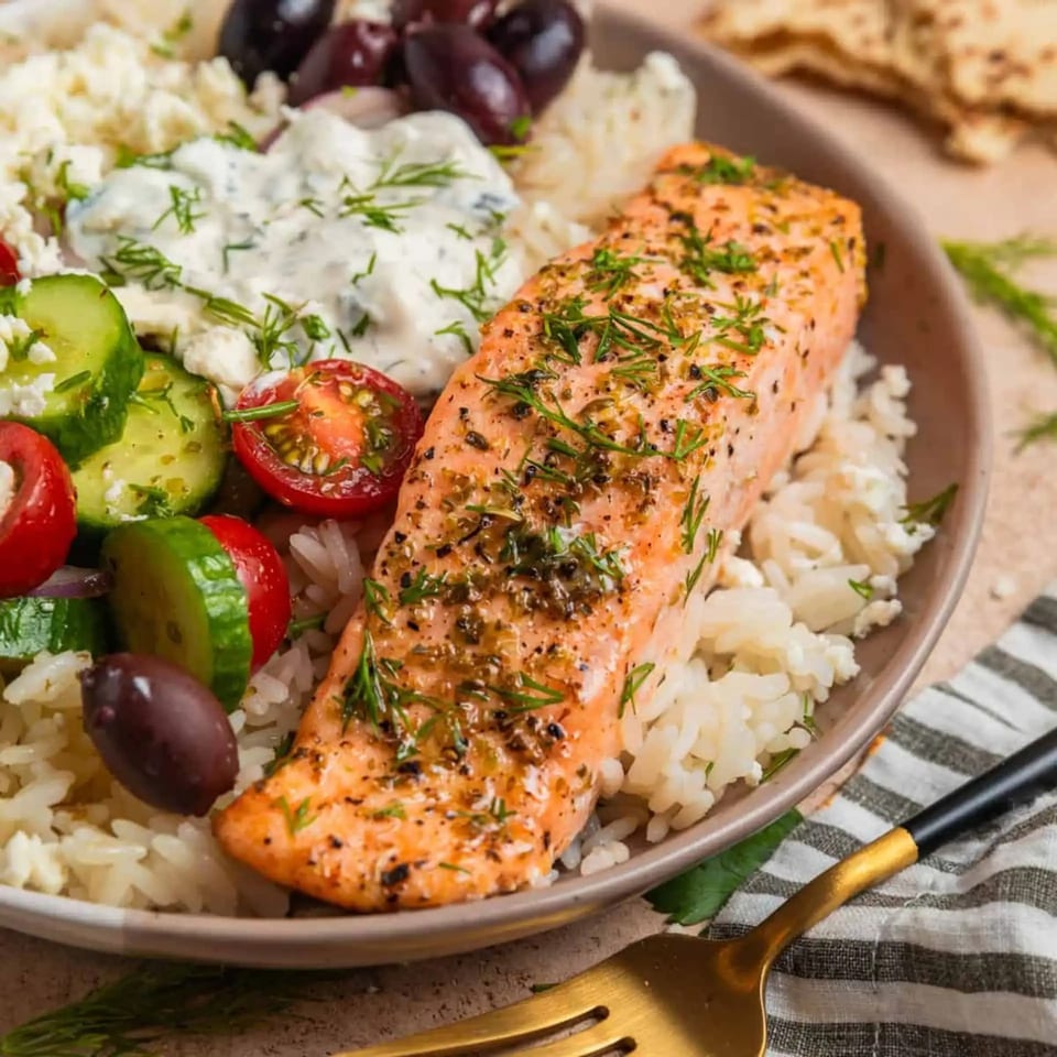 A close-up of a salmon fillet with dill, rice, tzatziki, cucumber, tomatoes, and olives.